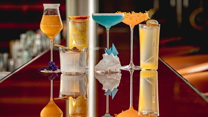 Line-up of six vibrant cocktails on a glossy bar counter, each uniquely presented with creative garnishes including flowers, citrus, a paper origami boat on cotton candy, and a flaming spice. The drinks vary in colour from golden amber and bright blue to soft peach and clear, reflecting elegantly on the polished surface.