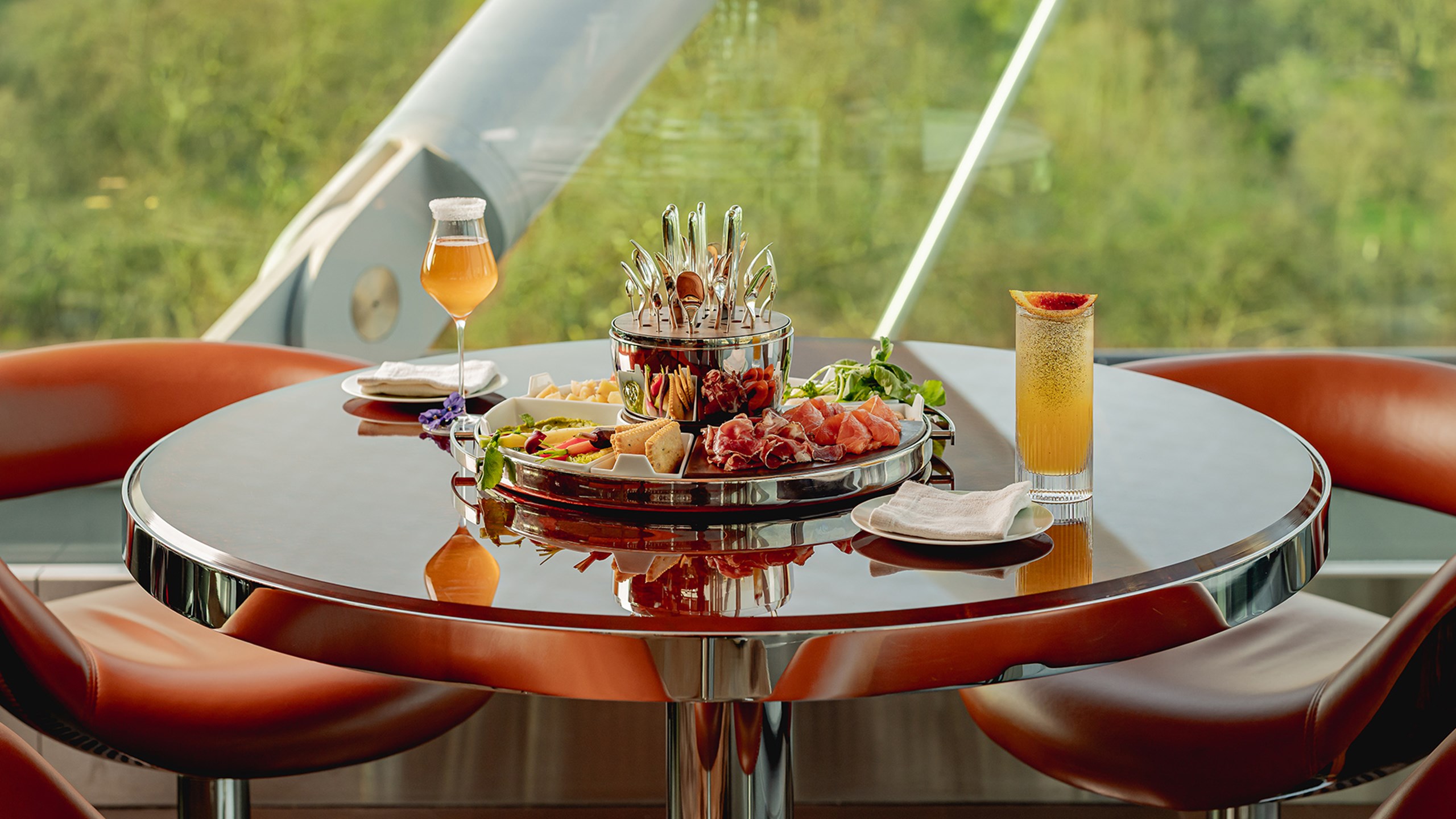 Round table with leather chairs featuring a platter of charcuterie, cheese, and crackers, served with two cocktails by large windows overlooking greenery.
