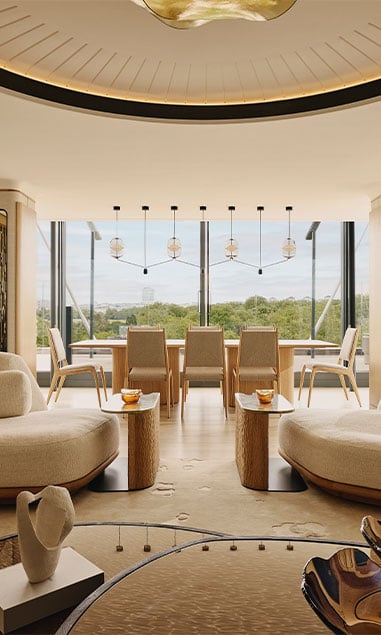 View of The Penthouse, with sofas and coffee tables, and in the background a dining table with chairs, and floor to ceiling windows overlooking Hyde Park.