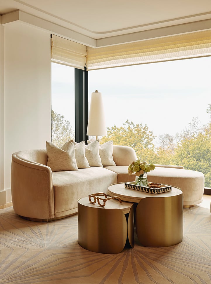 A serene living space with a plush curved sofa in soft neutral tones, complemented by sculptural brass tables and floor-to-ceiling windows offering a tranquil view of lush greenery.