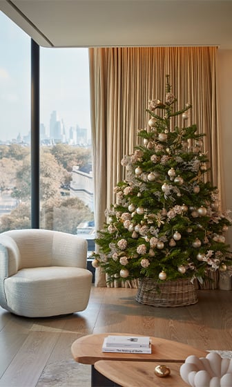 A beautifully decorated Christmas tree adorned with elegant gold and white ornaments stands in a modern, sunlit living room with a cozy armchair, a view of the city skyline, and warm, minimalist decor.