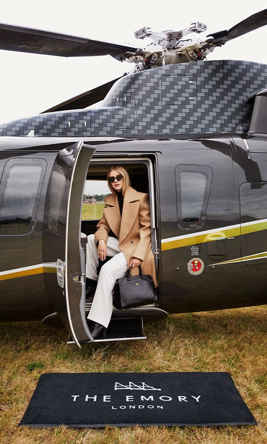 A stylish model woman is stepping out of a sleek helicopter, dressed in a beige overcoat and white pants, holding a black handbag. She is wearing sunglasses and standing on a mat that reads 