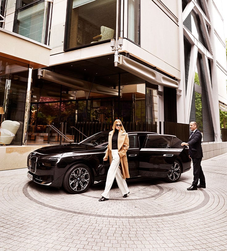 A stylish guest in a camel coat and sunglasses steps out of a sleek black car, as a doorman awaits in front of a modern, architecturally striking building.