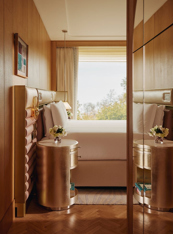 A luxurious bedroom with golden tones, featuring a plush bed, gold-accented nightstands, and soft natural light streaming through a large window.