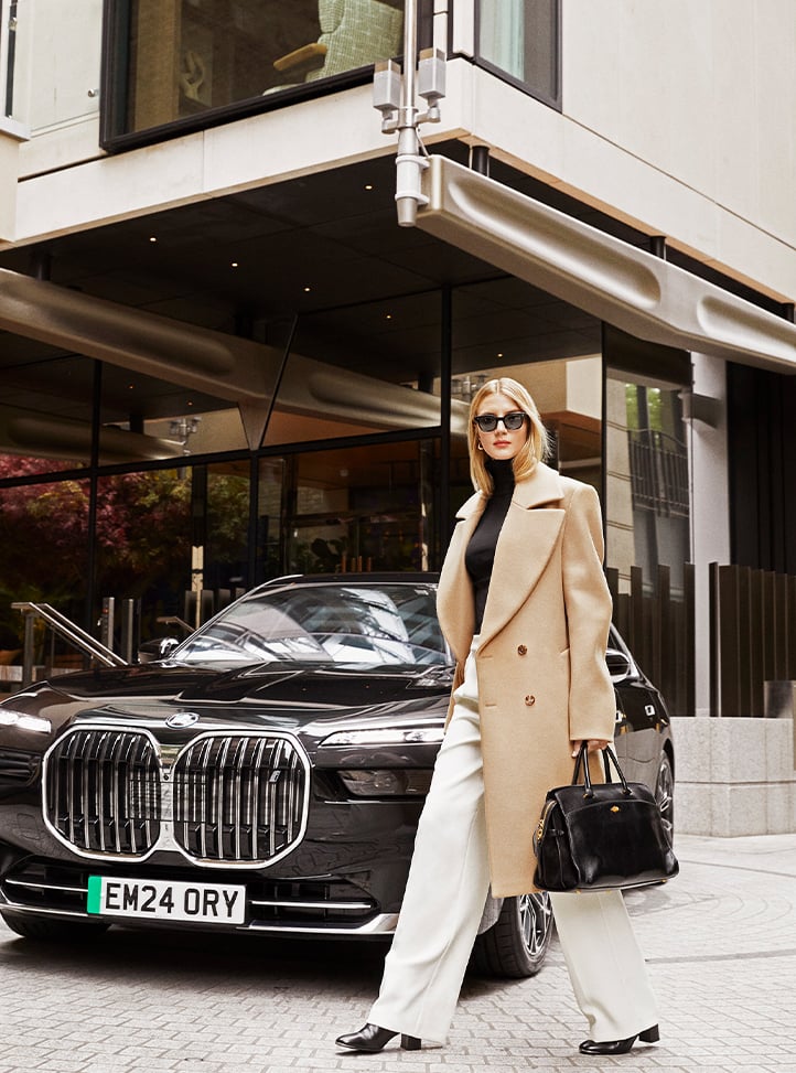 A fashionably dressed woman, carrying a black handbag, stands confidently in front of a luxury car and the contemporary façade of an upscale hotel.