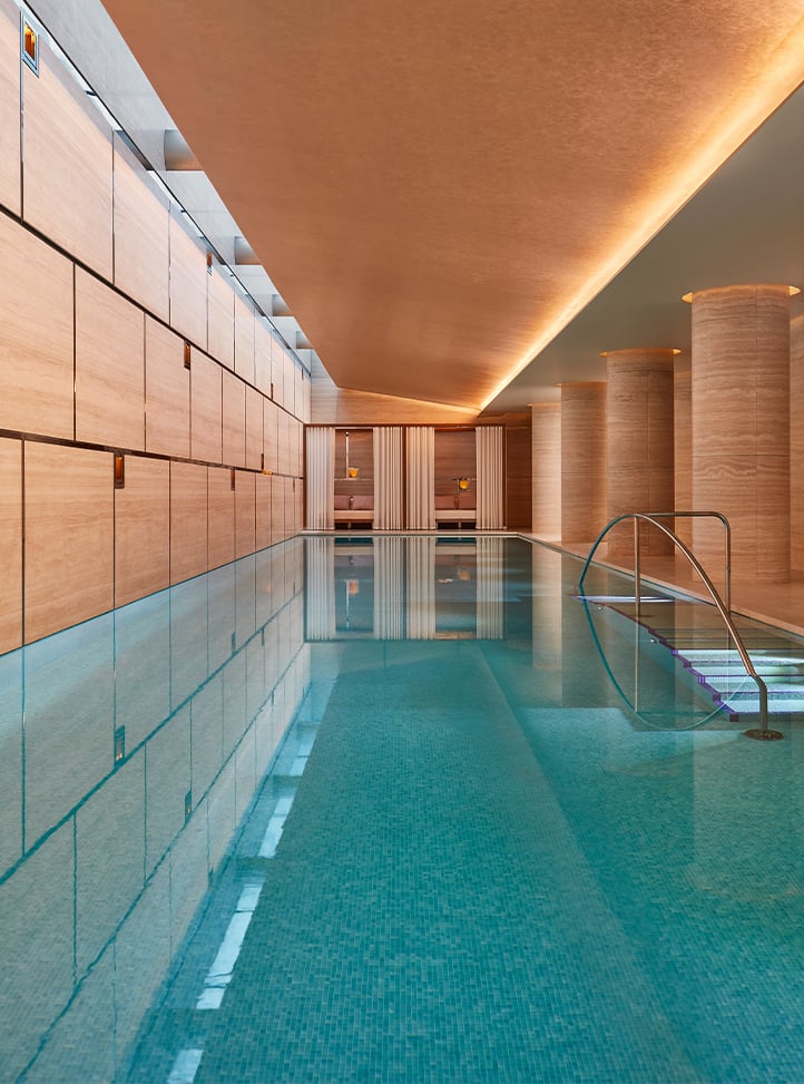 A serene indoor swimming pool with turquoise tiles, warm ambient lighting, and sleek architectural design featuring stone columns and soft drapery.