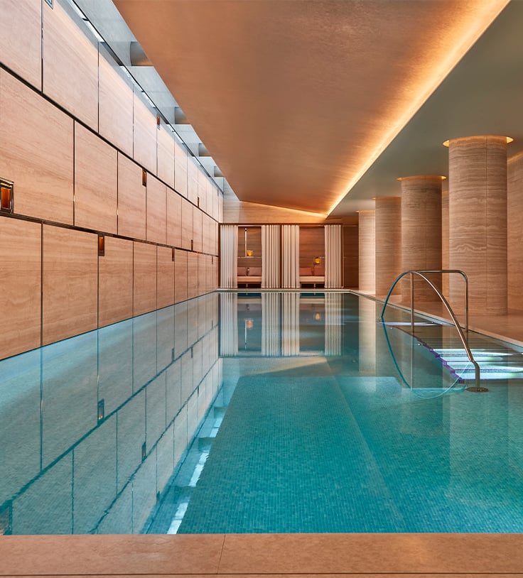 A sleek, light-filled swimming pool with tranquil waters, flanked by smooth stone walls and softly lit columns, offering a serene escape.