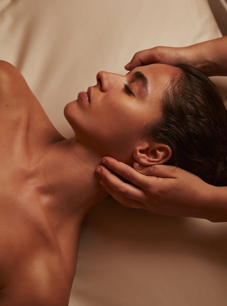 A woman with smooth, glowing skin receives a luxurious facial massage, her eyes closed in deep relaxation as skilled hands gently cradle her neck, set against a warm, serene backdrop.