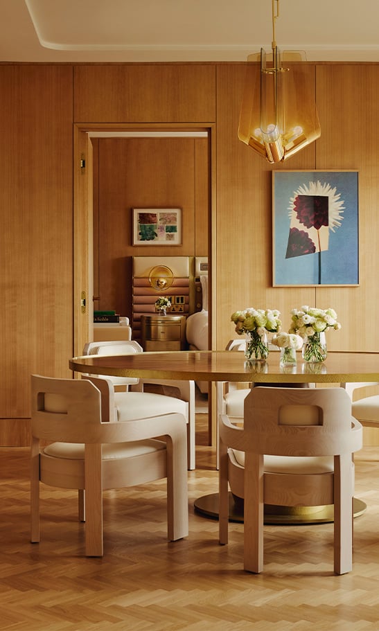 A modern suite dining area with a round wooden table, elegant beige chairs, and a large floral artwork on the wall, bathed in warm, natural light.