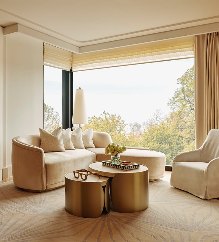 A cosy living room with a curving cream-coloured sofa, matching armchair, and sleek gold coffee tables, overlooking a scenic outdoor view through large windows.