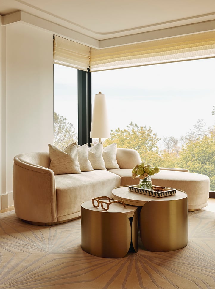 A cosy living room with a curving cream-coloured sofa, matching armchair, and sleek gold coffee tables, overlooking a scenic outdoor view through large windows.