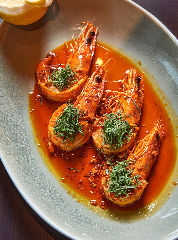 A beautifully plated dish featuring succulent prawns in a rich, red sauce, garnished with fresh herbs, served on a ceramic dish with a halved lemon on the side.