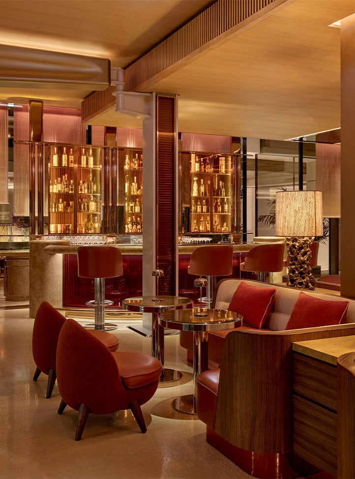 A stylish bar area with warm lighting, plush seating in shades of red and orange, and a backdrop of illuminated bottles displayed behind a polished bar.