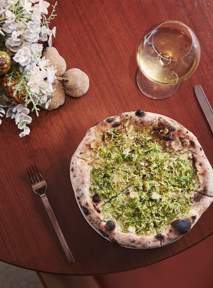 A festive table setting featuring a wood-fired pizza topped with shaved Brussels sprouts, accompanied by a glass of white wine and holiday-inspired decor.