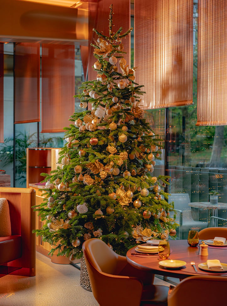 A grand Christmas tree adorned with golden ornaments and lights, set in a luxurious dining space with richly decorated tables and floor-to-ceiling windows.