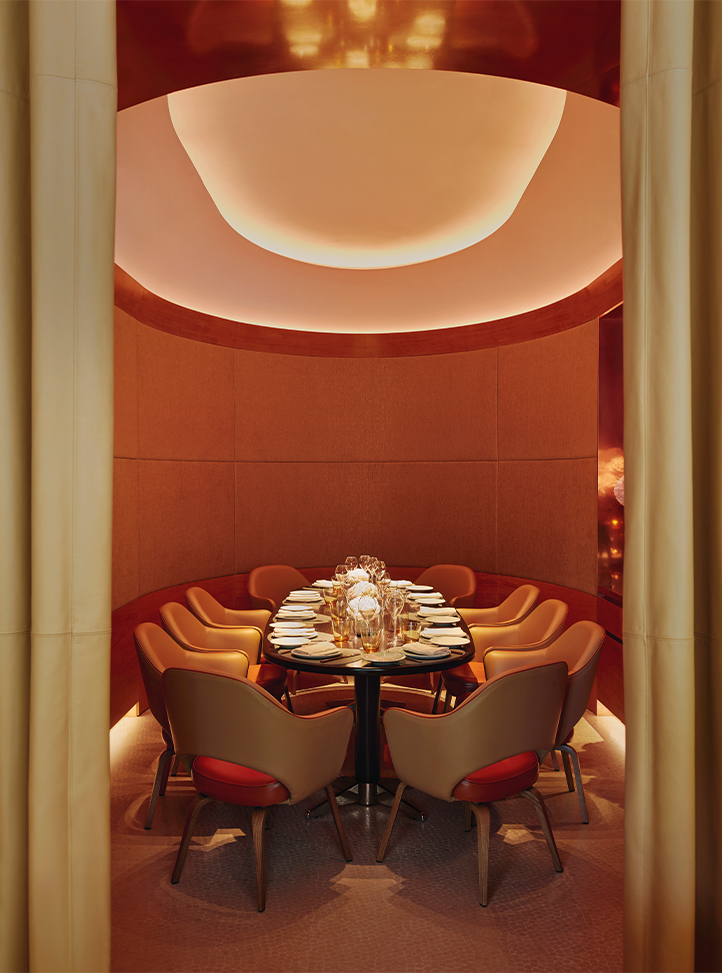 A luxurious, intimate private dining room with plush beige and red chairs arranged around a round table set for a sophisticated meal, bathed in warm, soft lighting from an elegantly designed ceiling.