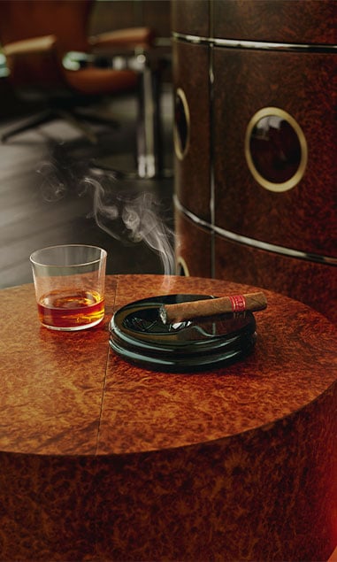 A small wooden table with a glass of whisky and a cigar that light, sitting on a black ashtray.