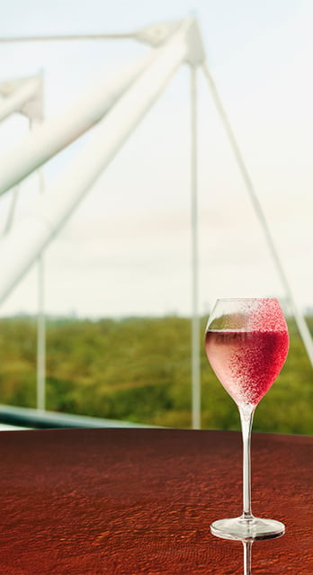 A stemmed glass filled with a pink beverage sits on a wooden table with a blurred outdoor scene, featuring large white structural elements and greenery, in the background.