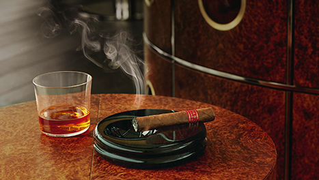 A cigar smoking on a tray, and a small glass of whisky sat on a dark red table in the Cigar Merchants