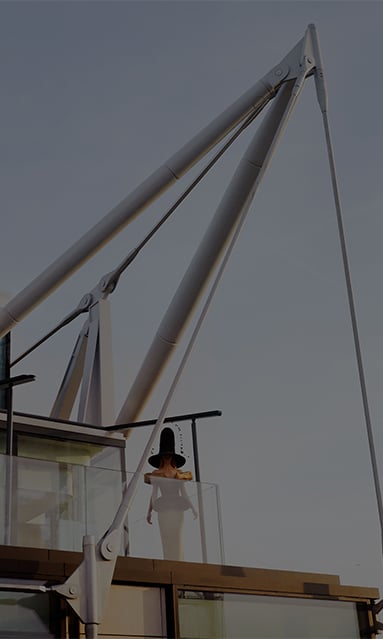 A woman wearing a tall, avant-garde black hat and a dramatic off-shoulder dress stands on a glass balcony surrounded by large industrial metal structures against a clear sky.