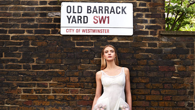 Model in white dress with brick wall behind her and street sign with 'Old Barrack Yard' on it