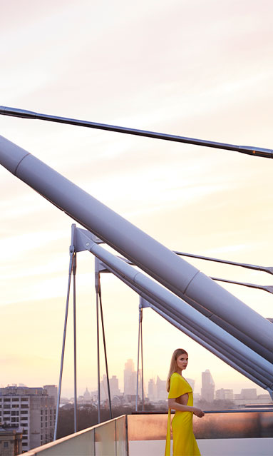 Model in yellow dress on The Emory Rooftop with architectural sails and London's urban landscape in the background.