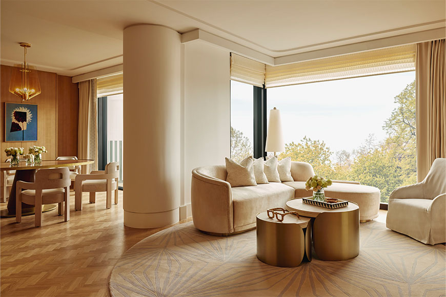 The Emory suite, golden and neutral colours with coffee table, vase of flowers and glasses, sofa and arm chair and floor to ceiling window behind, overlooking Hyde Park. In the backgroun on the left is dining table with chairs.