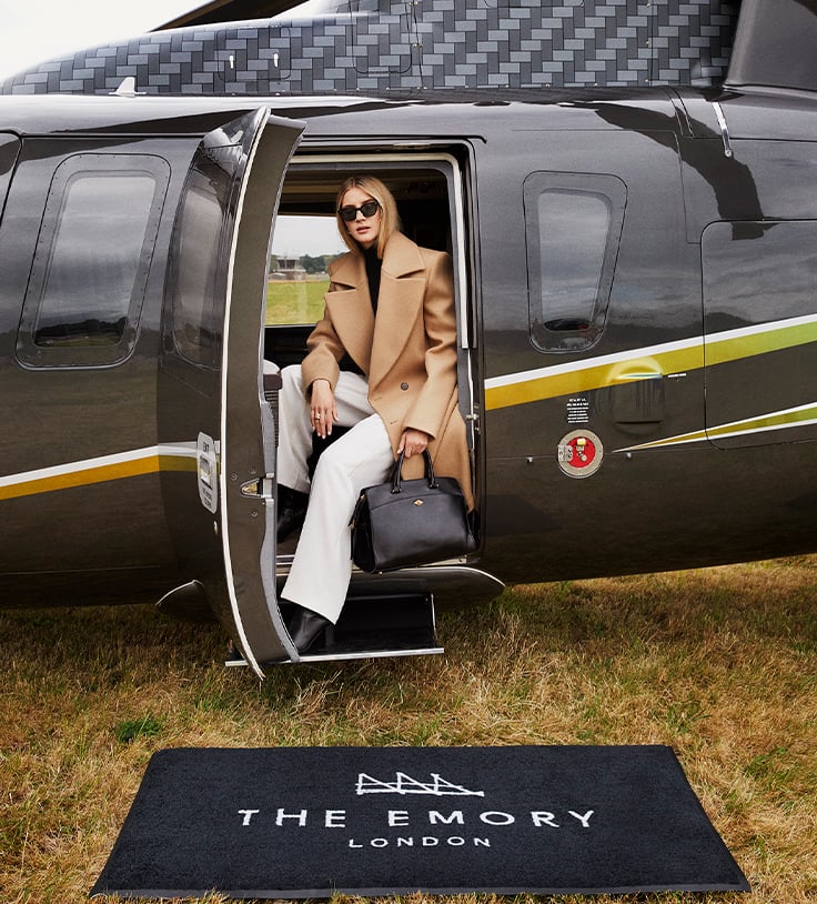 A stylish woman steps out of a sleek black helicopter onto a branded 'The Emory London' mat, embodying luxury travel.