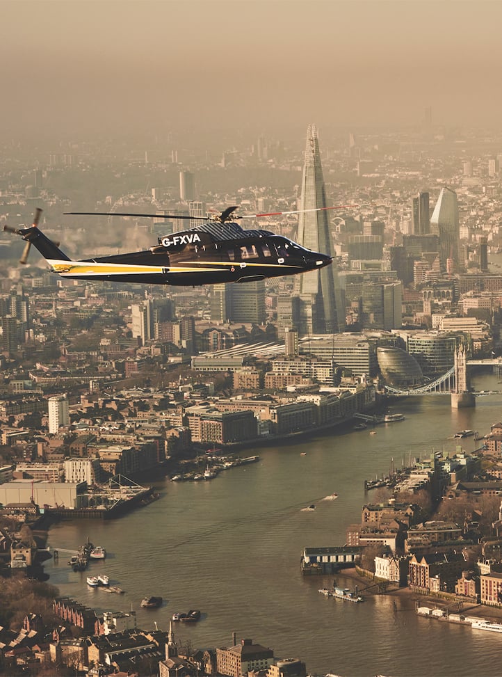 A sleek black and gold helicopter soars above London’s skyline, with The Shard and Tower Bridge visible through the hazy golden light of the cityscape.