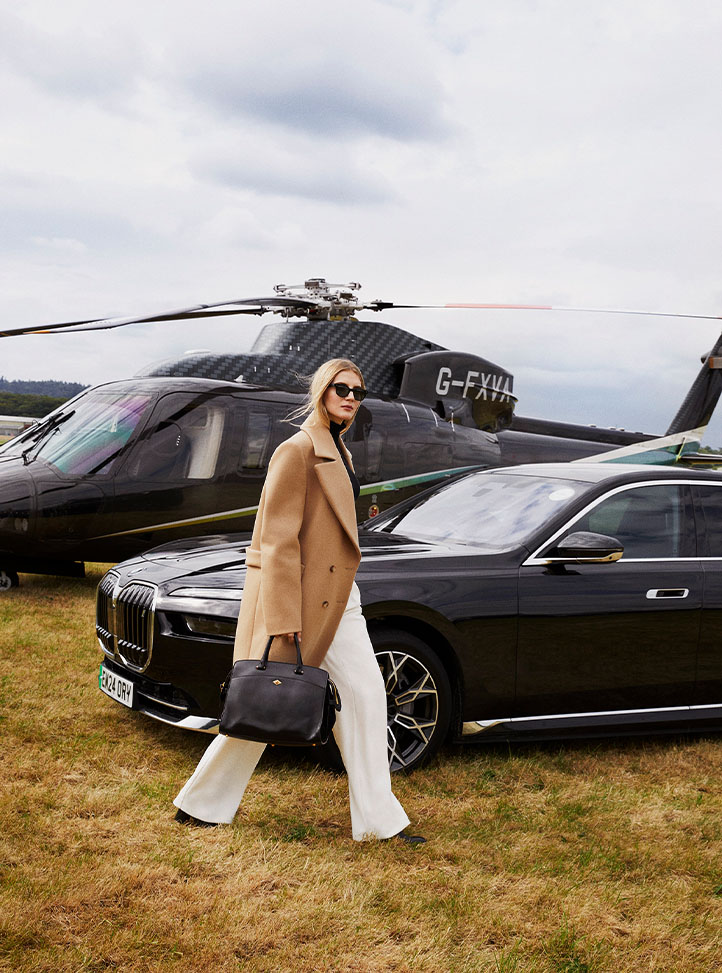 A sophisticated figure walks across a private airfield towards a chauffeured black BMW, with a state-of-the-art helicopter in the background.