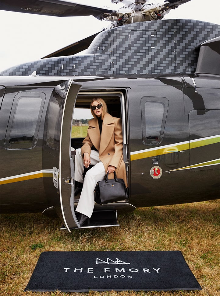 A poised traveler disembarks from a private helicopter, dressed in a camel coat and white trousers, carrying a black designer handbag.