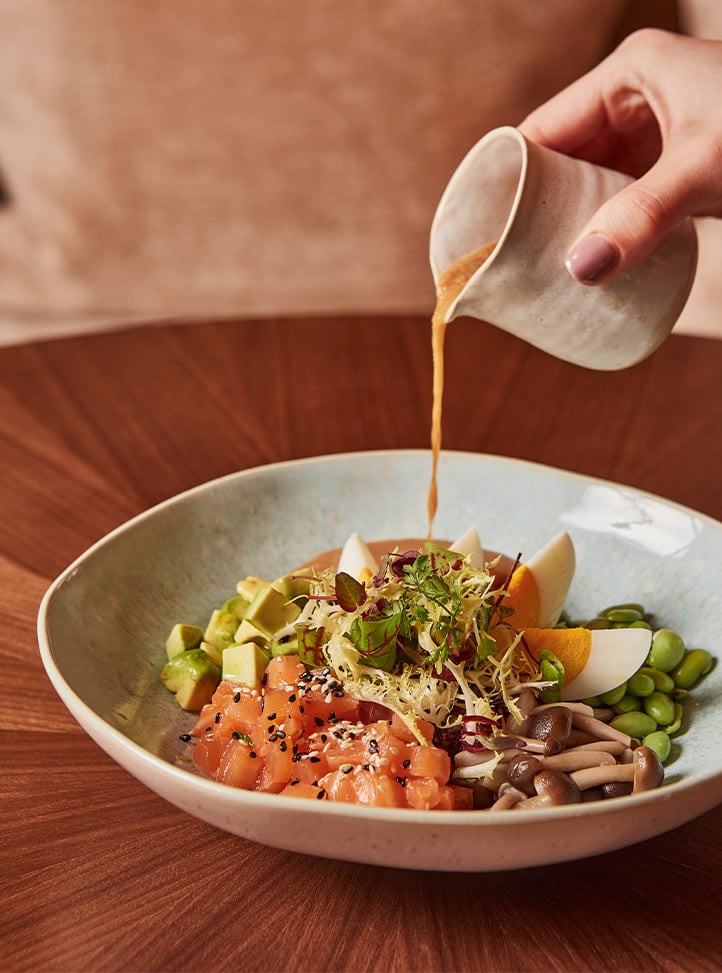 A hand gracefully pours a golden dressing over a vibrant poke-style salad with fresh salmon, avocado, eggs, mushrooms, and greens, elegantly presented in a ceramic bowl.