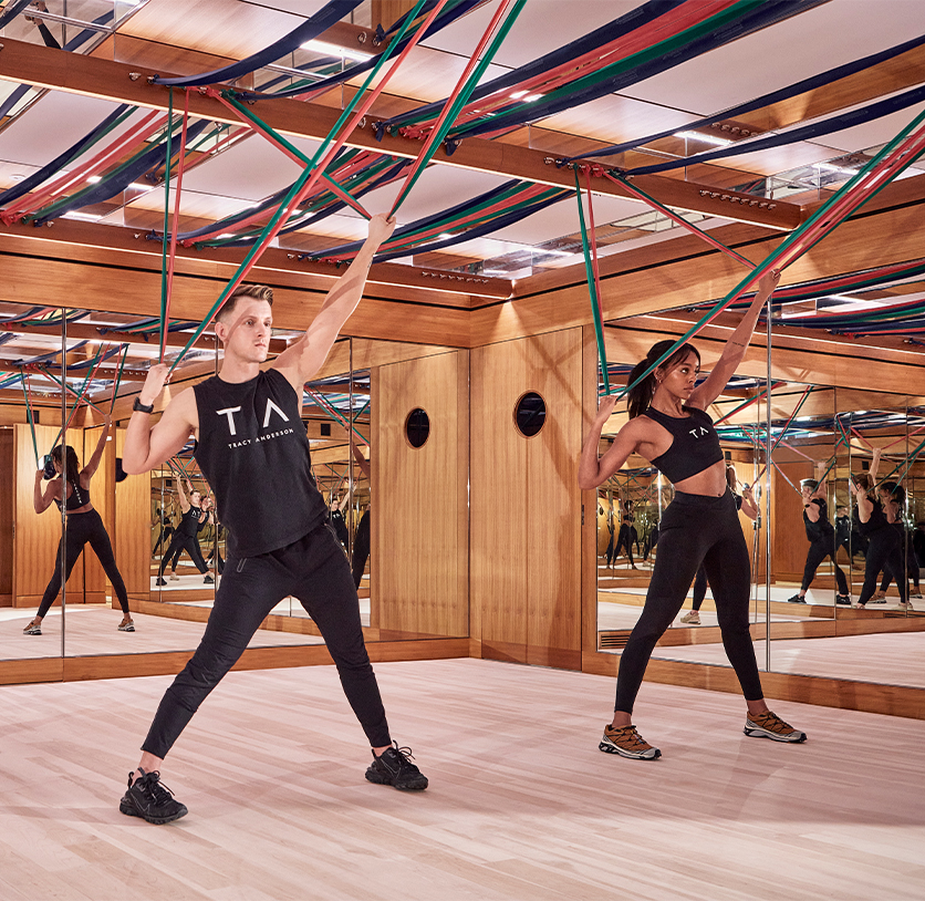 Two individuals in black Tracy Anderson workout gear engage in a resistance band workout in a mirrored studio with wooden paneling and colorful ceiling straps.