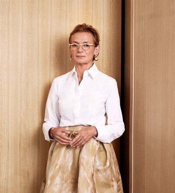 A distinguished woman in a crisp white blouse and a flowing gold skirt stands against warm wooden paneling, exuding sophistication with timeless simplicity.