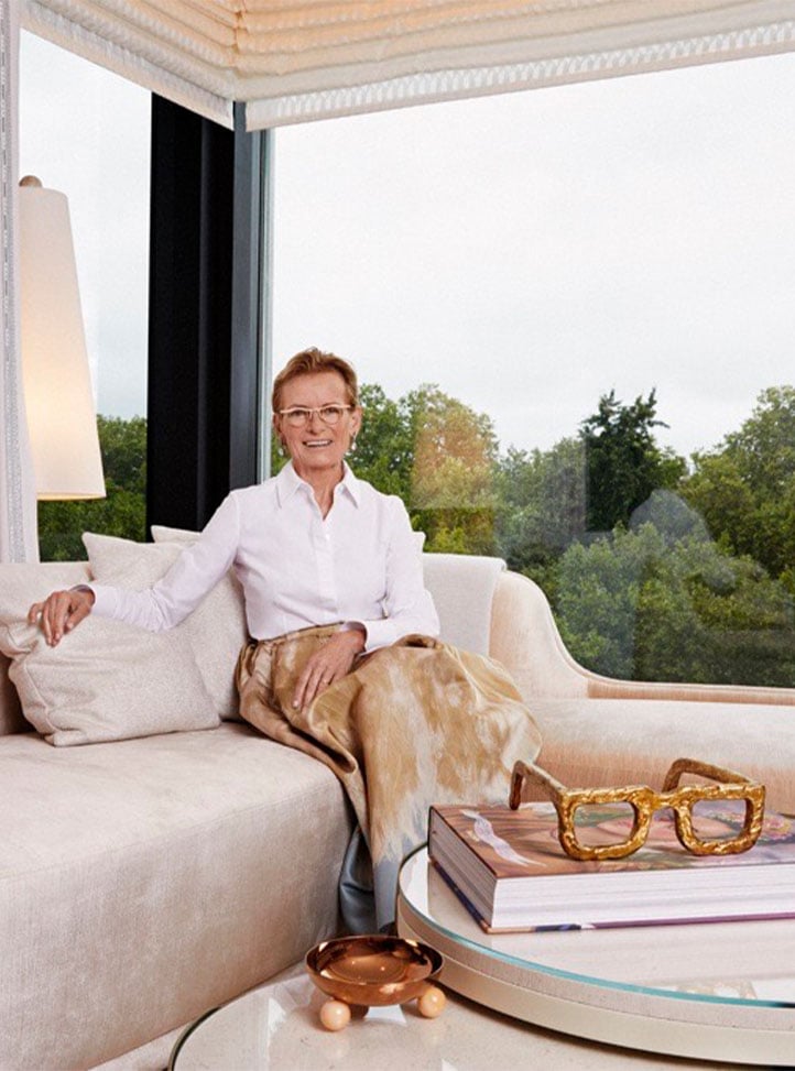 A distinguished woman in a crisp white blouse and a flowing gold-toned skirt sits gracefully on a plush cream sofa in a sunlit space with floor-to-ceiling windows overlooking lush greenery, surrounded by elegant décor.