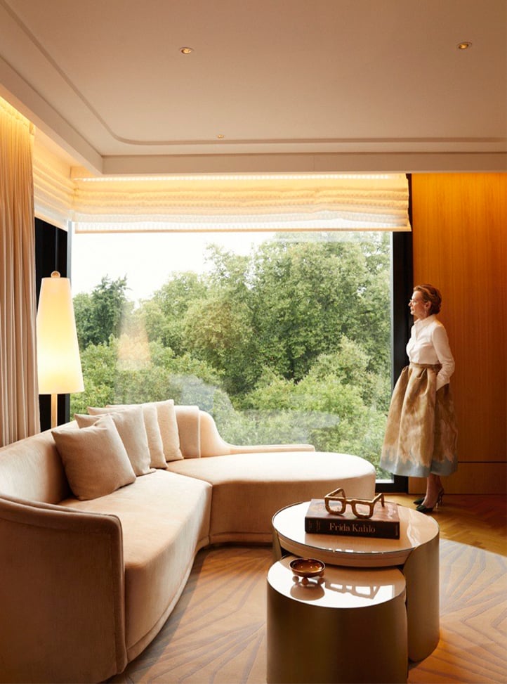 A poised woman in a white blouse and gold skirt stands by a floor-to-ceiling window, overlooking lush greenery, in an elegantly designed living space with a sculptural sofa and curated decor.