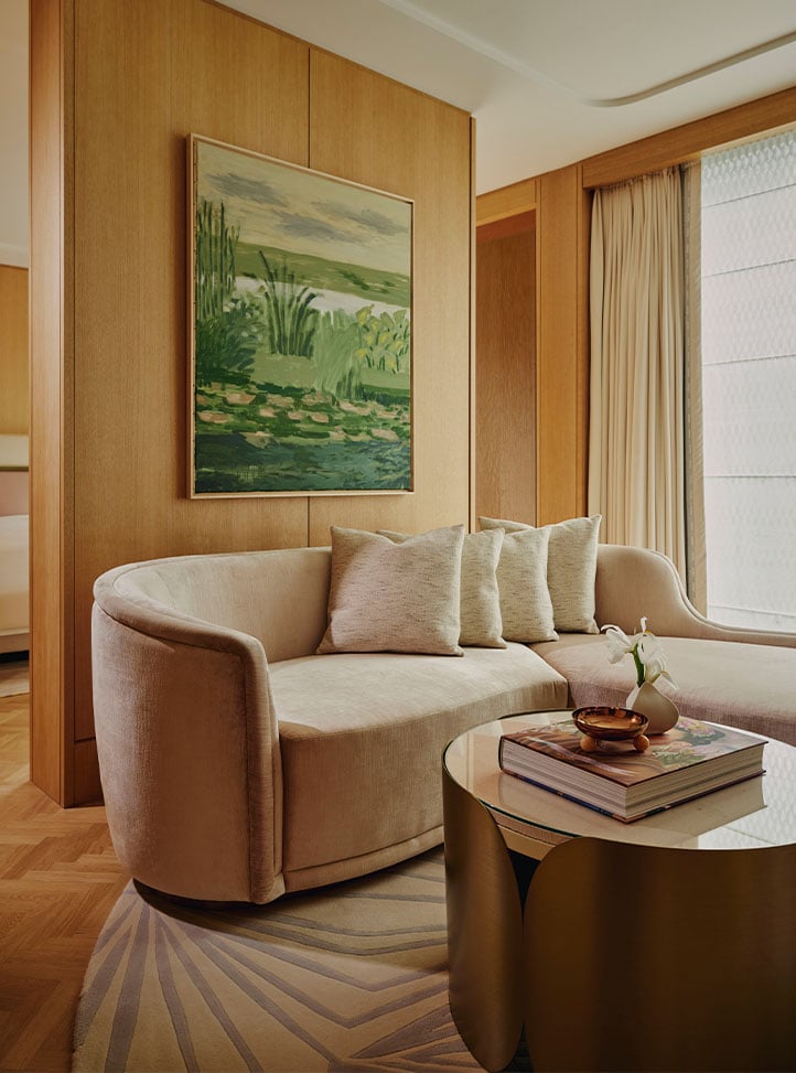 A serene living space featuring a curved cream sofa adorned with textured cushions, a sculptural gold coffee table, and a large abstract green landscape painting against warm wooden paneling, bathed in soft natural light.