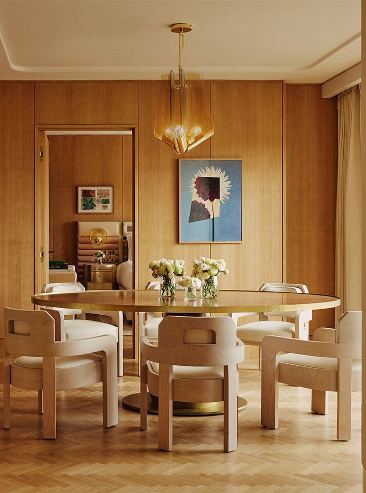 An elegant dining space with a round gold table surrounded by minimalist sculpted chairs, illuminated by a modern amber-toned pendant light, with curated artwork and fresh floral arrangements enhancing the sophisticated ambiance.