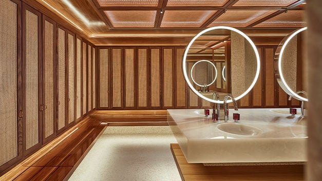 A luxurious washroom featuring warm wood paneling, ambient lighting, and a central vanity with illuminated circular mirrors, exuding a refined elegance