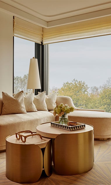 Corner of pale pink sofa, sitting in front of glass windows revealing leafy green trees outside. In the forefront is a gold rounded coffee table with stylish coffee table books and a gold pair of statement glasses as decor on top.