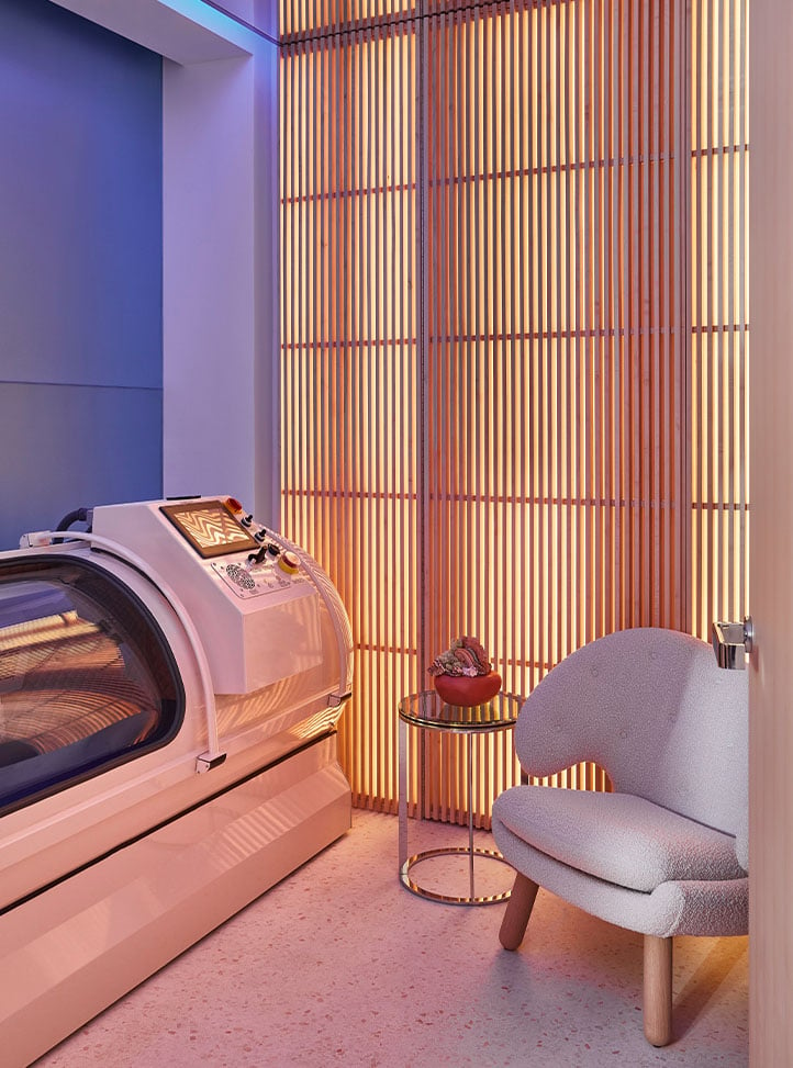 A tranquil wellness space featuring a sleek hyperbaric chamber, warm wood paneling, and a plush lounge chair with soft lighting.