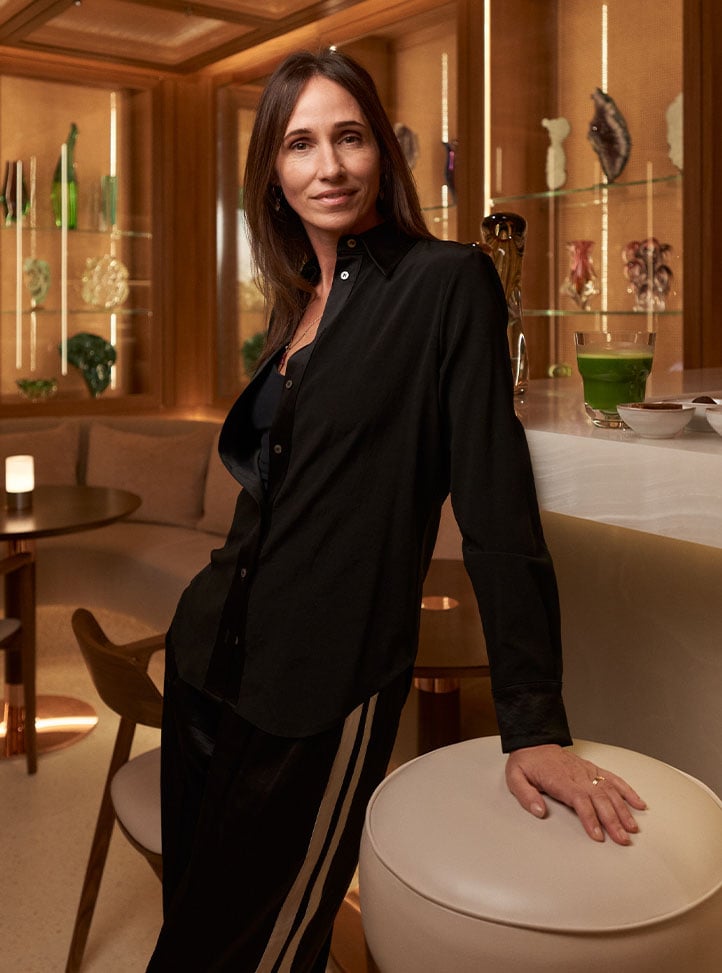 A poised woman in an elegant black outfit leaning on a sophisticated bar, set against a backdrop of curated glass art and warm, ambient lighting.